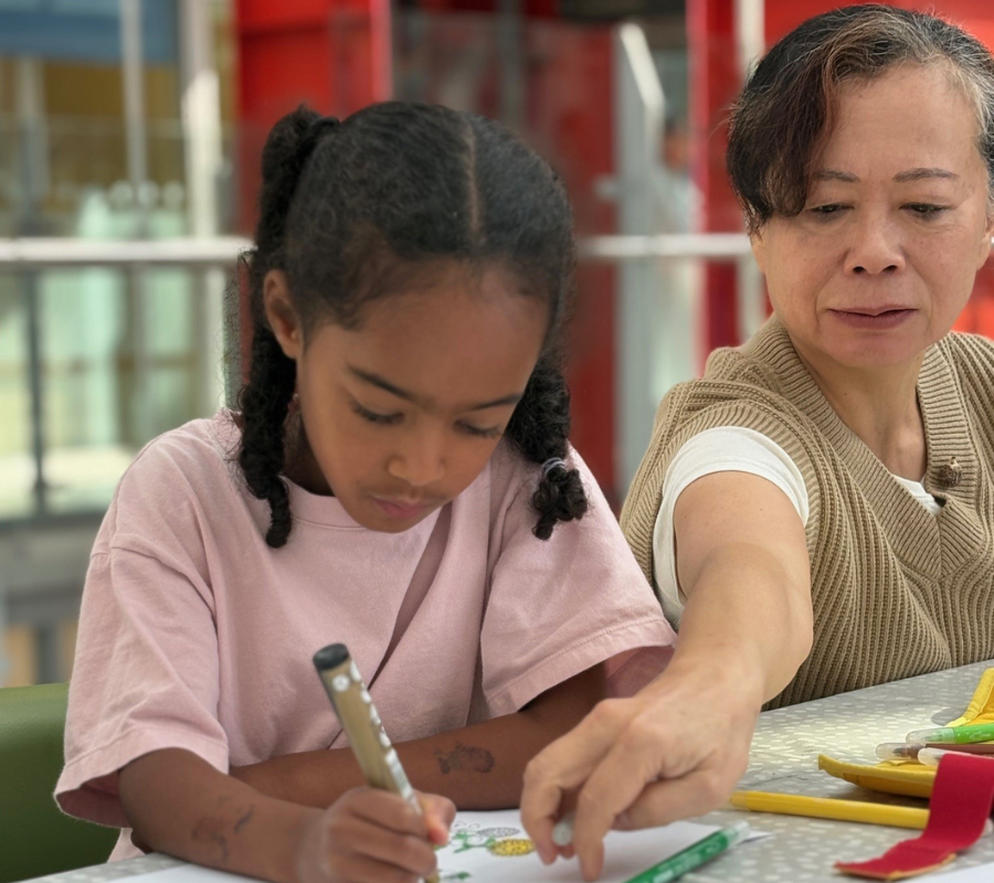 A child and Kiriko sitting at a table, the child is drawing on a sheet of paper and Kiriko is pointing to it