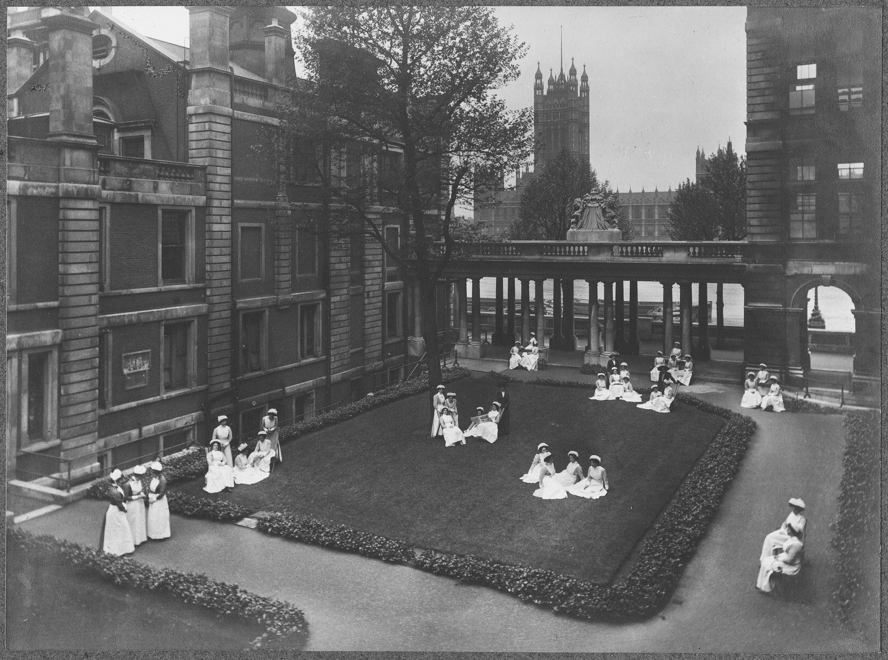 A black and white photo of nurses sitting on a grass area between hospital buildings. They are wearing full length nursing uniforms and aprons and hats